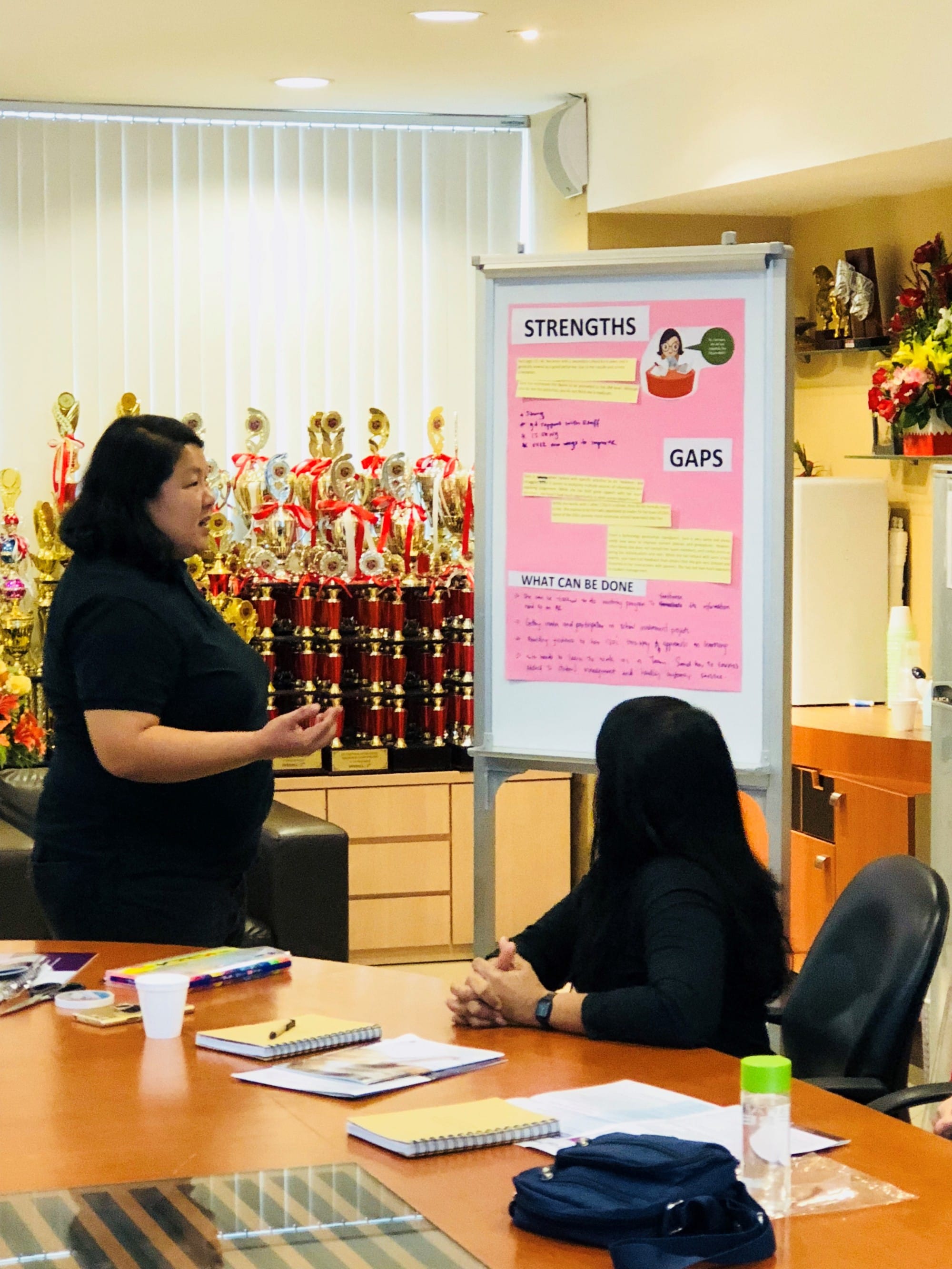 Woman presenting beside a pink poster board labeled Strengths, Gaps, and What Can Be Done to a seated attendee.